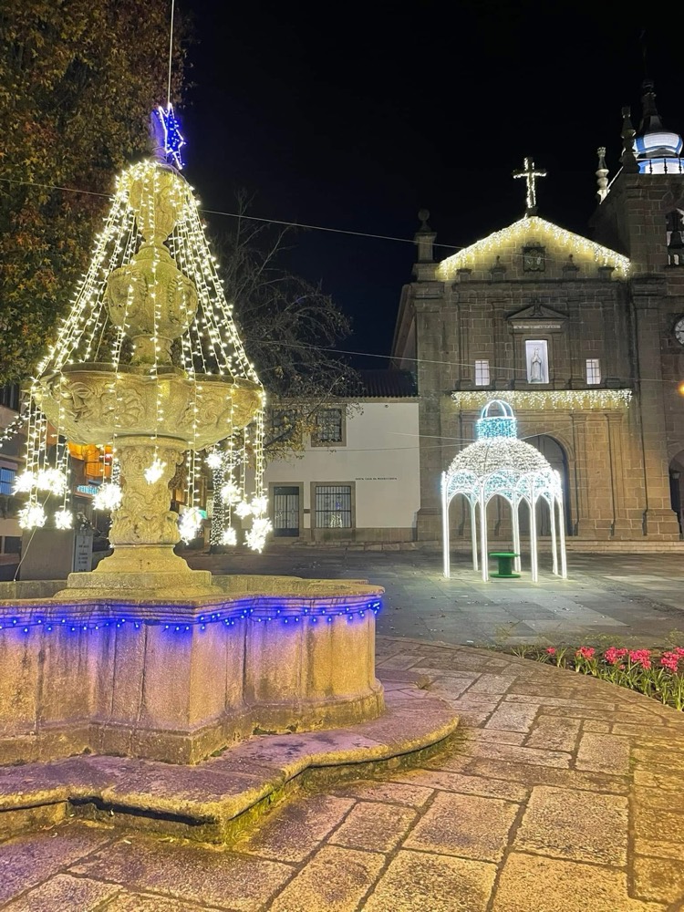 Chafariz de pedra iluminado com cordões de luz e coreto luminoso em frente a uma igreja